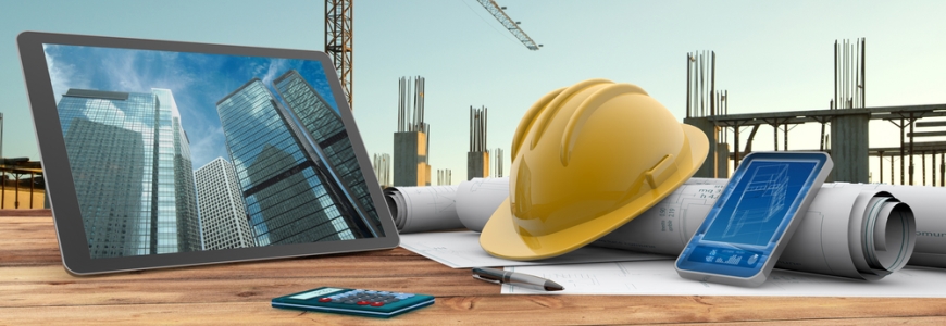 Tablet showing office buildings on a table filled with construction plans, a hard hat, calculator and documents. In the background is the start of the construction of the buildings