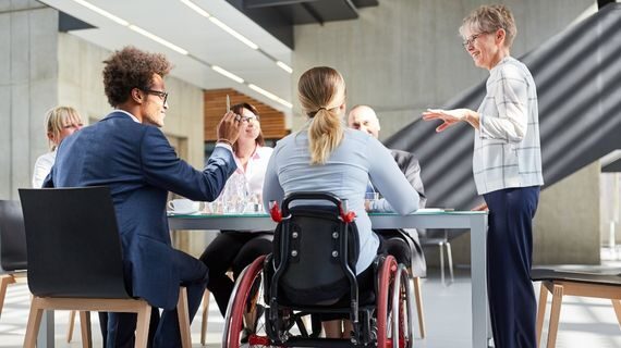 Six colleagues around a table. Four are sitting in chairs and one is in a wheelchair and one is standing.