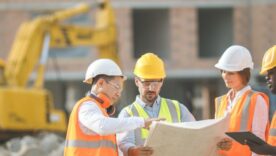 Male and female builders, architect and engineers with draft plan of building and laptop computer talking on constructing site