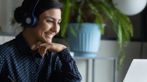 Head shot happy young indian ethnicity female manager wearing wireless headphones, looking at laptop screen, holding pleasant conversation with partners clients online, working remotely at workplace.