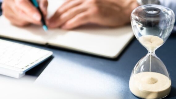 Close-up Of Hourglass In Front Of Businessperson's Hand Calculating Invoice.