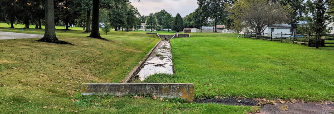 Water Drainage Easement with Water in Ditch and Green Grass on All Sides and Small Patch of Asphalt Blacktop and Trees in the Distance.