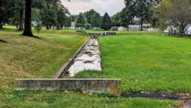 Water Drainage Easement with Water in Ditch and Green Grass on All Sides and Small Patch of Asphalt Blacktop and Trees in the Distance.