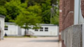 Red-brick school building corner with white trim and gray stone accent - pavement, school portable classrooms, trees in background.