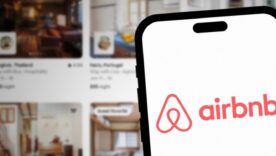 Airbnb logo on smartphone screen with Airbnb website and rooms in background.