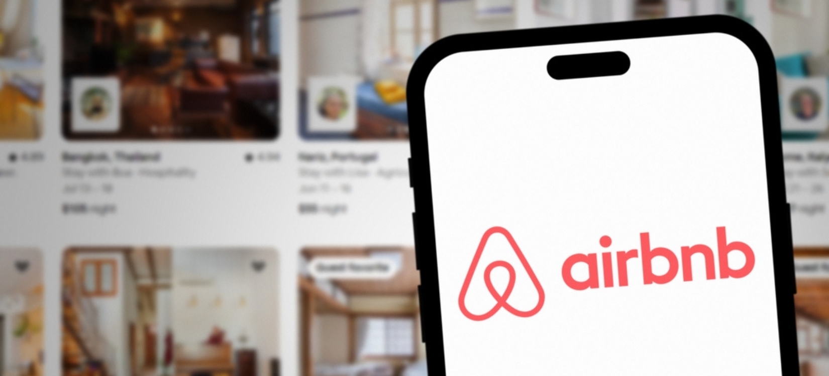 Airbnb logo on smartphone screen with Airbnb website and rooms in background.